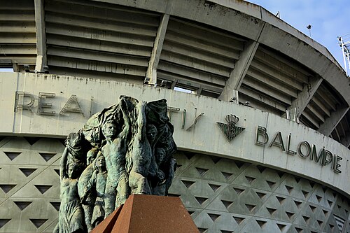 Vista de la fachada principal del Estadio Benito Villamarín, en primer plano un monumento en bronce situado en la esquina entre Avenida de la Palmera y…